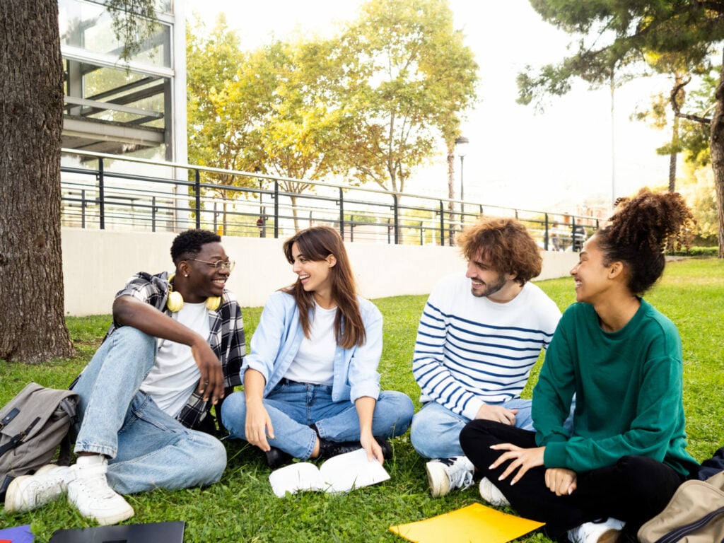 Group of diverse college student friends laughing and chatting sitting on the grass on campus. High quality photo