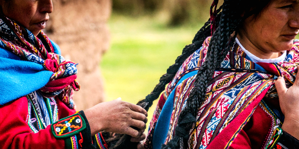 A Peruvian woman braiding another Peruvian woman's hair, both dressed in traditional Peruvian clothing.